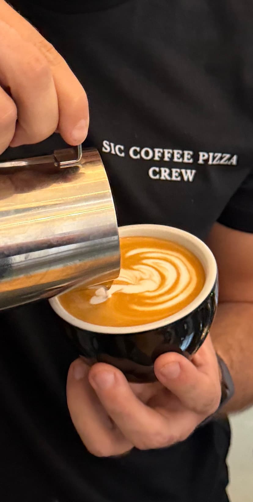 Barista im SIC Coffee Pizza Crew-Shirt gießt Latte Art