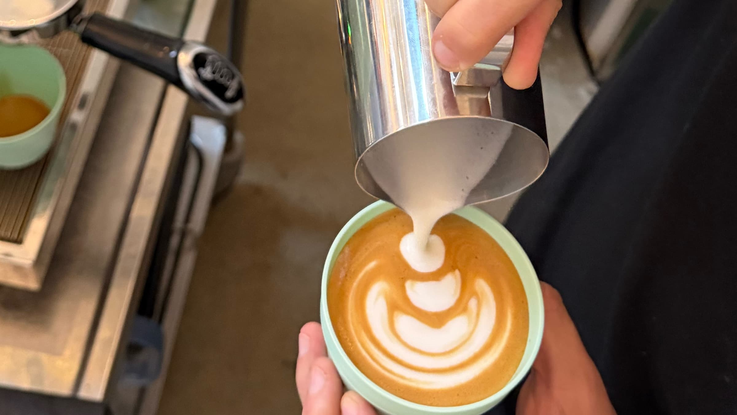 Nahaufnahme: Milchschaum läuft aus dem Kännchen in einen Cappuccino und formt Latte Art