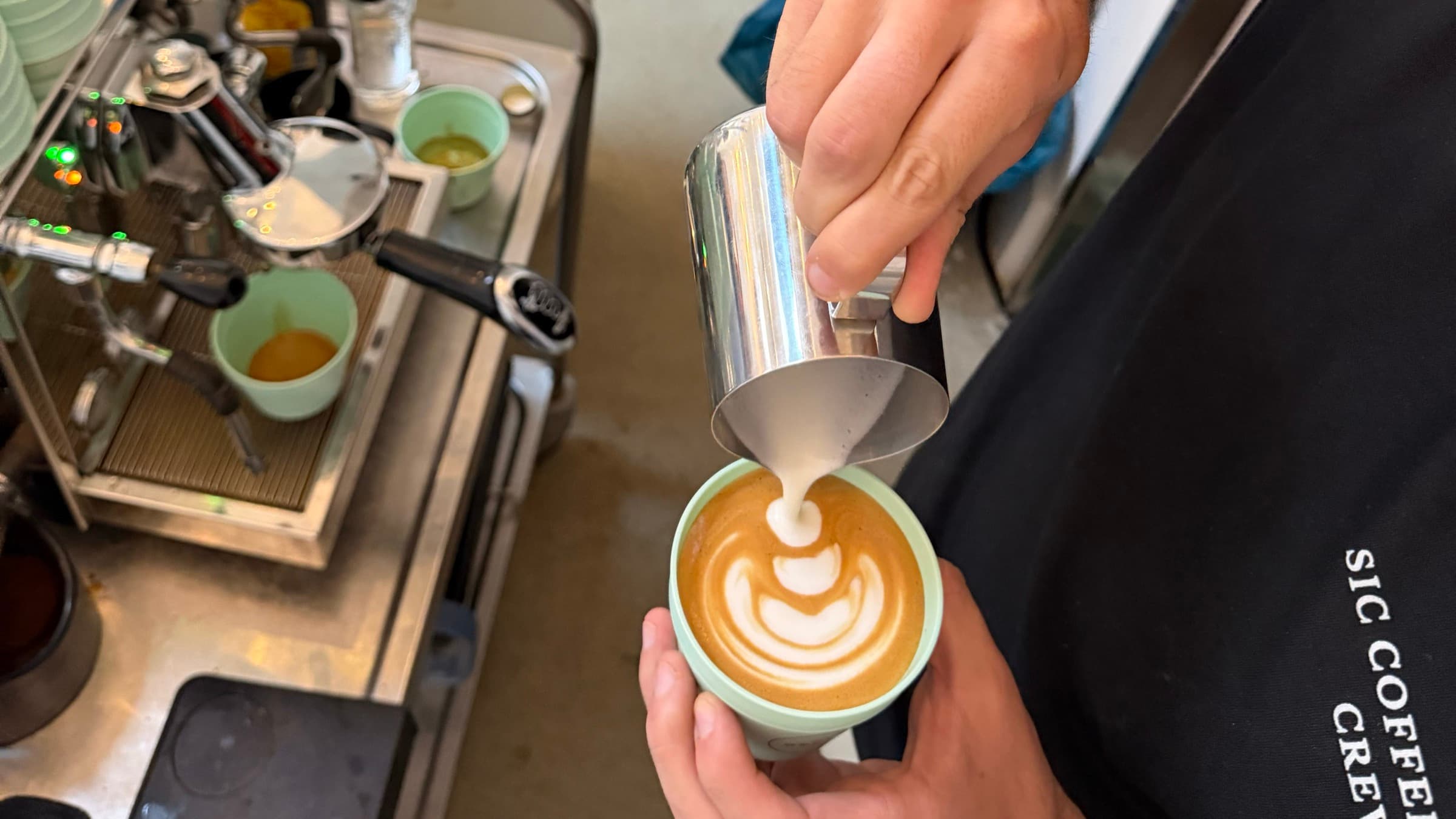 Barista gießt Latte Art in einen Becher, im Hintergrund die Siebträgermaschine